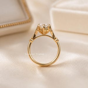 May include: A gold engagement ring with a large, clear gemstone. The ring features an intricate, detailed setting with decorative elements. The band is smooth and polished. The ring is displayed on a soft, cream-colored surface.
