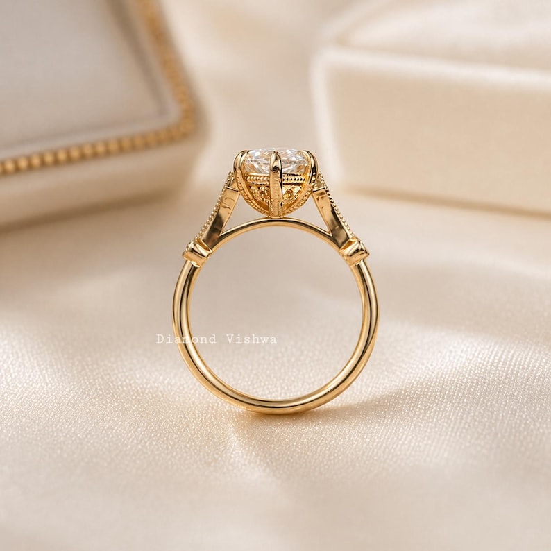 May include: A gold engagement ring with a large, clear gemstone. The ring features an intricate, detailed setting with decorative elements. The band is smooth and polished. The ring is displayed on a soft, cream-colored surface.