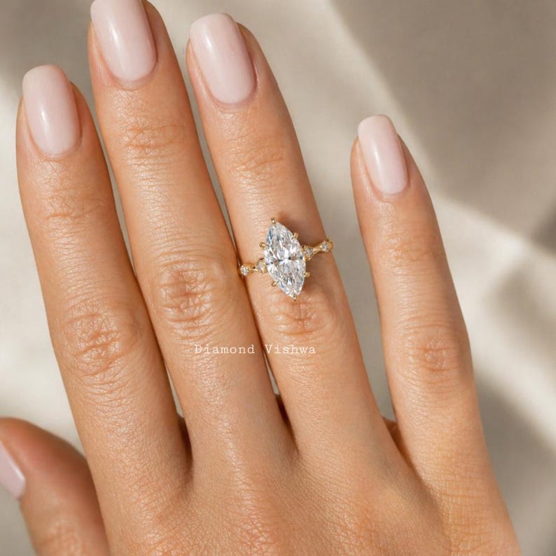 May include: A marquise-cut diamond engagement ring with a gold band. The ring features a large, elongated diamond as the centerpiece, with smaller accent diamonds on the band. The ring is set on a hand with manicured nails.