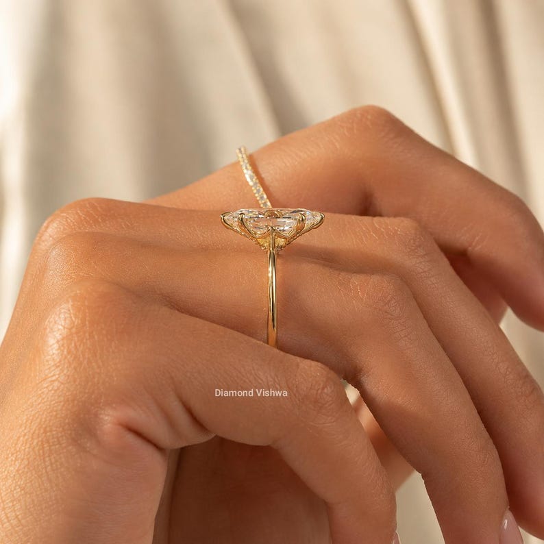May include: A gold ring featuring a large, oval-cut diamond. The ring's band is slender, and the diamond is set with delicate prongs. The ring is held up against a soft, neutral background. The text "Diamond Vishwa" is visible.