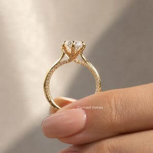 May include: A gold engagement ring with a large, clear gemstone. The band is adorned with small, sparkling stones. The ring is held by a person with a light skin tone. The text "Diamond Vishwa" is visible.