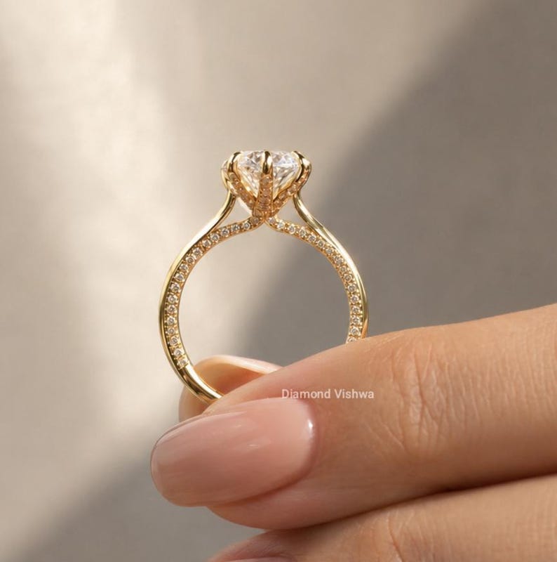May include: A gold engagement ring with a large, clear gemstone. The band is adorned with small, sparkling stones. The ring is held by a person with a light skin tone. The text "Diamond Vishwa" is visible.