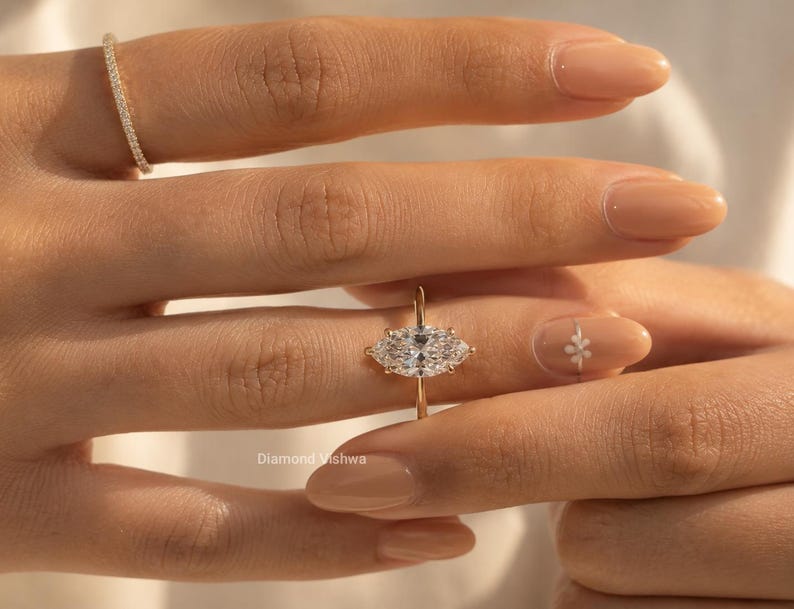 May include: A close-up of a hand wearing a gold ring with a large, oval-shaped diamond center stone and two smaller side stones. A smaller gold band with small diamonds is on the adjacent finger. The ring is set against a background of a person's skin.