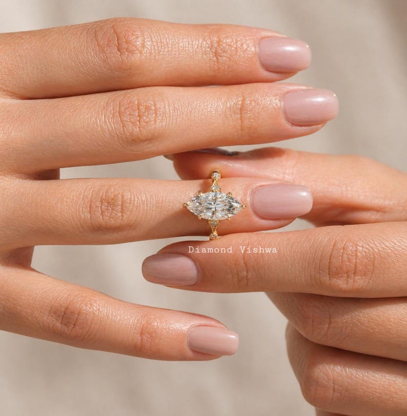 May include: A gold ring with a marquise-cut diamond center stone and smaller accent diamonds. The ring is held between two hands. The text "Diamond Vishwa" is visible on the ring.