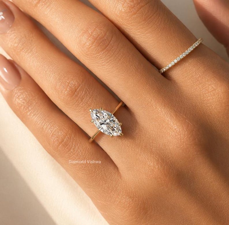 May include: A gold ring with a marquise-cut diamond is displayed on a hand. The diamond is the focal point, reflecting light. A second, smaller ring with small diamonds is also visible. The text "Diamond Vishwa" is at the bottom.
