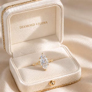 May include: A marquise-cut diamond ring in an open, cream-colored jewelry box. The ring has a gold band with small accent diamonds. The box is lined with cream-colored velvet and has the text "DIAMOND VISHWA" printed inside.