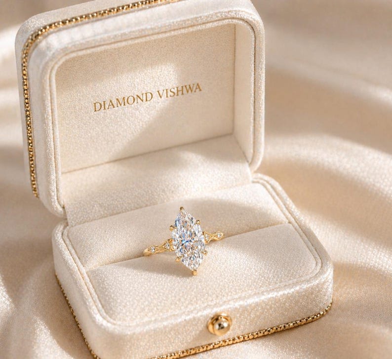 May include: A marquise-cut diamond ring in an open, cream-colored jewelry box. The ring has a gold band with small accent diamonds. The box is lined with cream-colored velvet and has the text "DIAMOND VISHWA" printed inside.