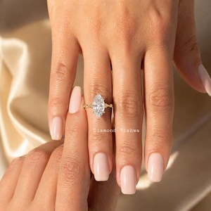 May include: A marquise-cut diamond engagement ring with a gold band. The ring is set on a hand with manicured nails. The ring has a large, clear diamond as the focal point. The band has small accent stones.