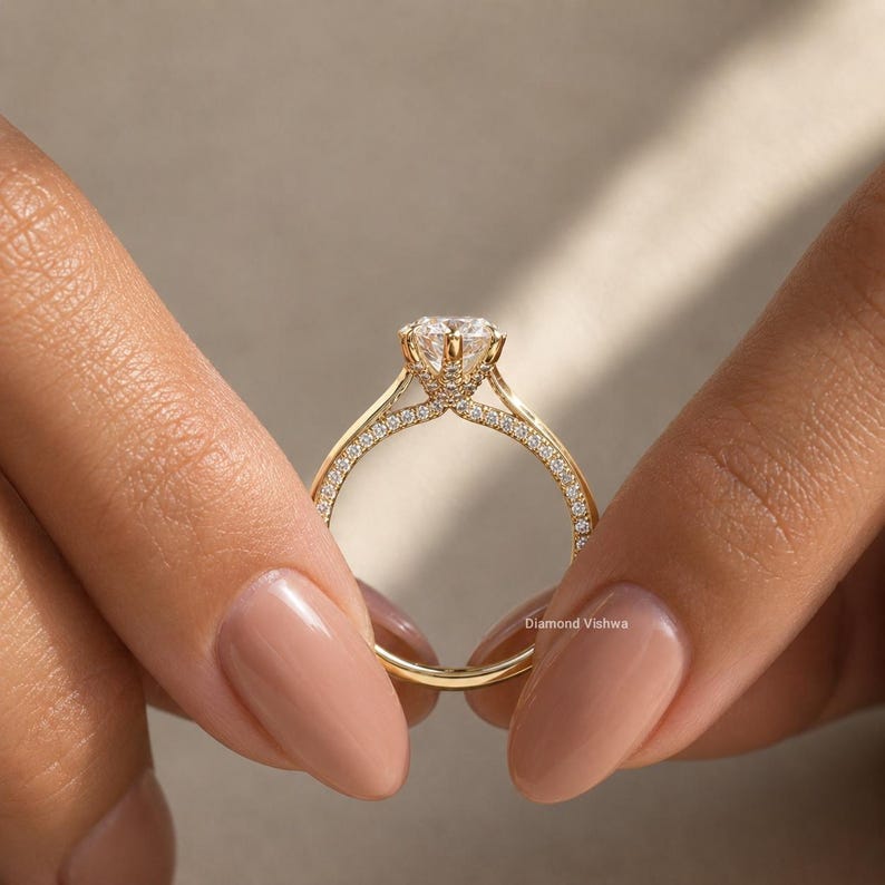 May include: A gold ring with a large, clear gemstone at the center. The band is adorned with small, sparkling stones. The ring is held between two hands with manicured nails. The ring is a piece of jewelry.