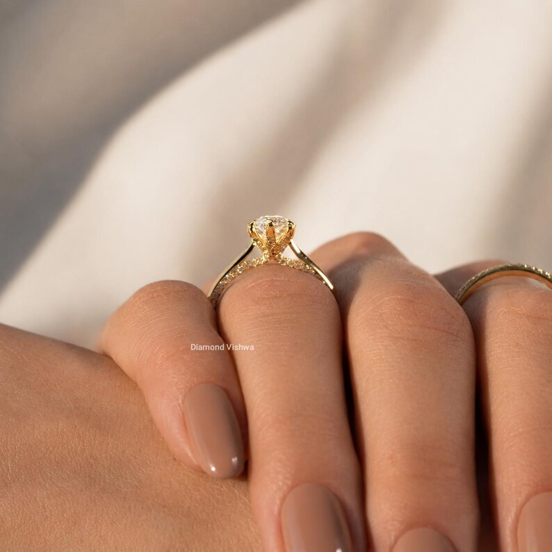 May include: A gold engagement ring with a large, round diamond center stone. The band is also adorned with smaller diamonds. The ring is on a person's finger. The text "Diamond Vishwa" is visible.