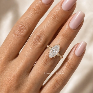 May include: A gold engagement ring featuring a marquise-cut diamond. The ring has a delicate band with small accent stones. The ring is displayed on a hand with manicured nails. The text "Diamond Vishwa" is visible.