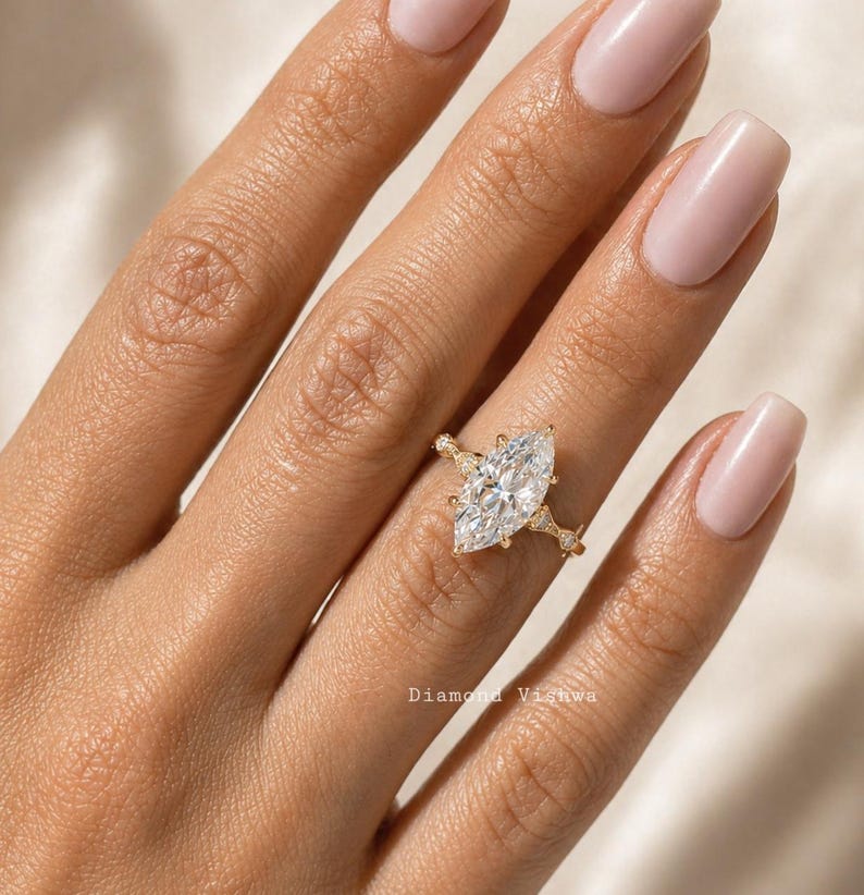 May include: A gold engagement ring featuring a marquise-cut diamond. The ring has a delicate band with small accent stones. The ring is displayed on a hand with manicured nails. The text "Diamond Vishwa" is visible.