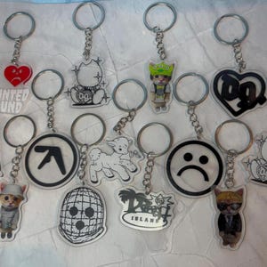 May include: A collection of thirteen acrylic keychains featuring various designs including a red heart with the text I HAUNTED MOUND, a black and white cat, a cat in a green hat, a black heart with DG, a person, a cat in a white hat, an Aphex Twin logo, a white lamb, a black and white mask, a Drain Island logo, a sad face, a cat in a suit, and a white cat.