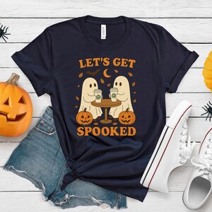 May include: Dark blue t-shirt featuring the phrase "LET'S GET SPOOKED" and a graphic of two ghost figures at a table with coffee cups, pumpkins, and a crescent moon. Halloween-themed decorations surround the shirt.