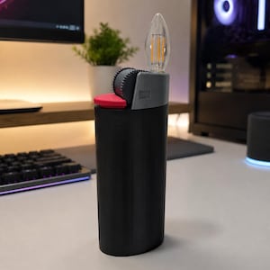 May include: A black cylindrical lighter-shaped object featuring a grey top section with a textured ignition wheel, a small red button, and a clear, flame-shaped LED filament bulb extending from the top, mimicking the appearance of a lit flame.
