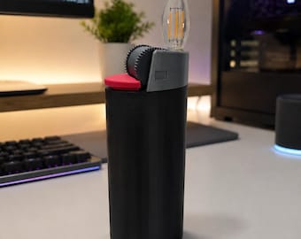 Könnte beinhalten: Ein schwarzes, zylindrisches Objekt in Feuerzeugform mit einem grauen Oberteil, einem strukturierten Zündrad, einem kleinen roten Knopf und einer klaren, flammenförmigen LED-Filamentbirne, die oben herausragt und eine Flamme imitiert.