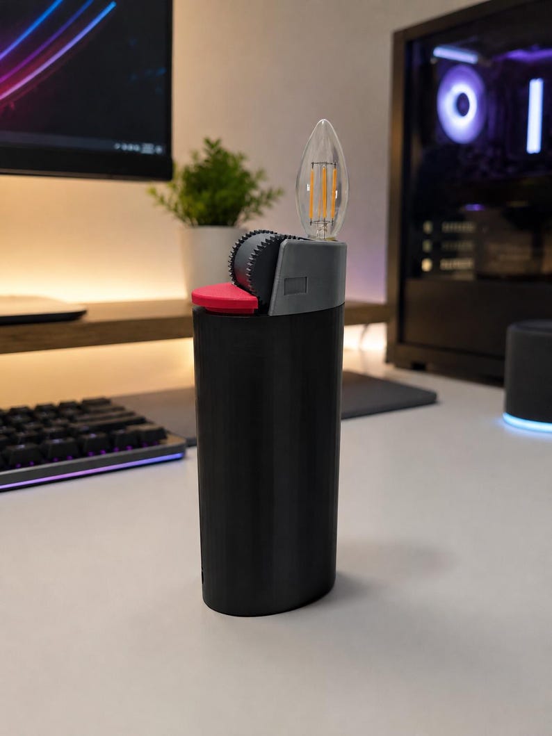 May include: A black cylindrical lighter-shaped object featuring a grey top section with a textured ignition wheel, a small red button, and a clear, flame-shaped LED filament bulb extending from the top, mimicking the appearance of a lit flame.