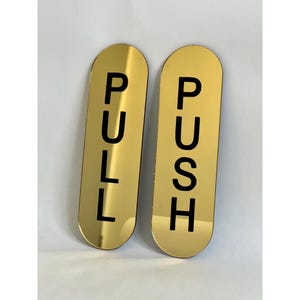Puede incluir: Dos letreros dorados para puertas con extremos redondeados. Un letrero dice "PULL" en letras mayúsculas negras, y el otro dice "PUSH" en letras mayúsculas negras. Los letreros son probablemente para un espacio comercial o público.