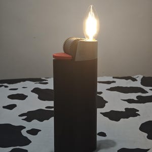 May include: A dark brown rectangular lighter-shaped lamp with a red ignition button and a grey serrated wheel. A glowing, flame-shaped LED filament bulb is positioned at the top where the flame would typically emerge, emitting a warm light.