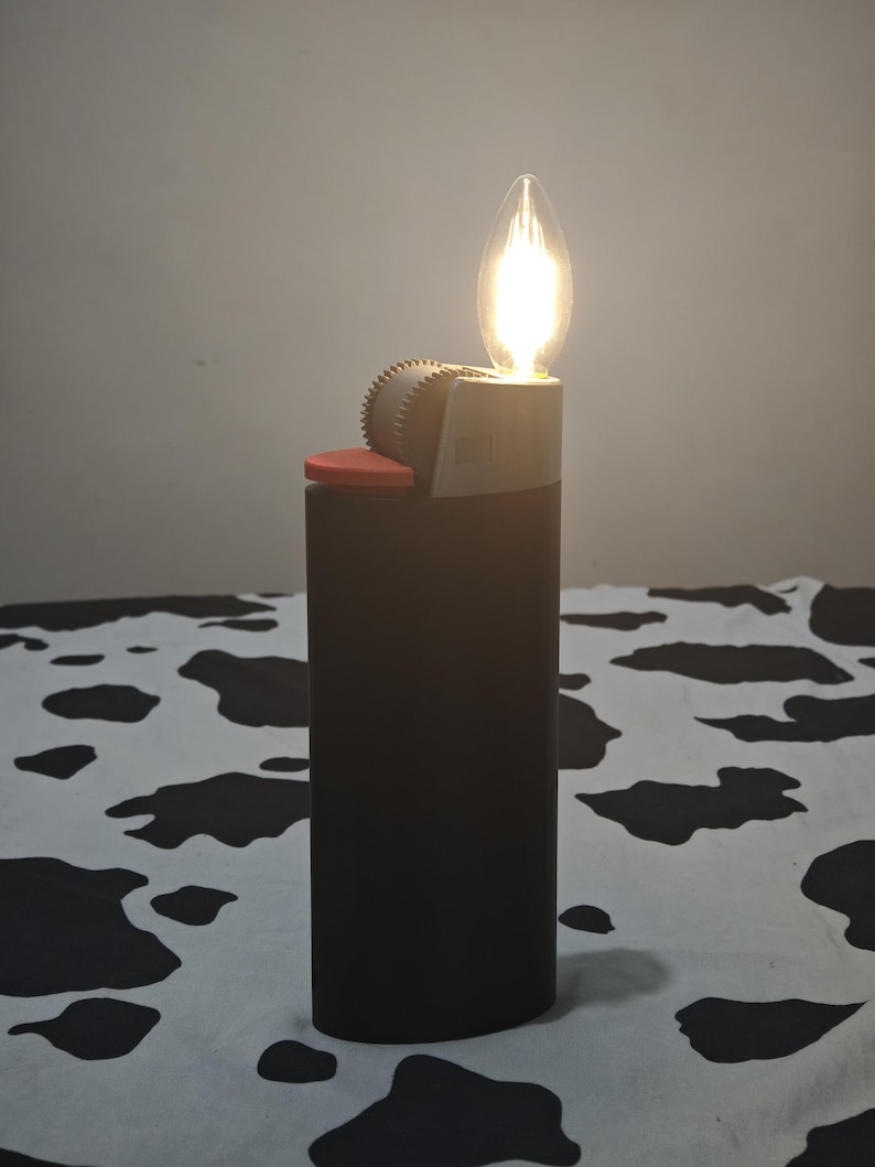 May include: A dark brown rectangular lighter-shaped lamp with a red ignition button and a grey serrated wheel. A glowing, flame-shaped LED filament bulb is positioned at the top where the flame would typically emerge, emitting a warm light.