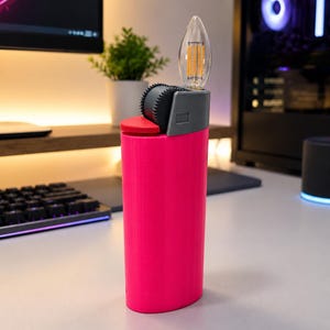 May include: A bright pink cylindrical lighter-shaped lamp featuring a grey top section with a textured ignition wheel and a clear, flame-shaped LED filament bulb extending from the top. The base is solid pink with a smooth, vertical finish.