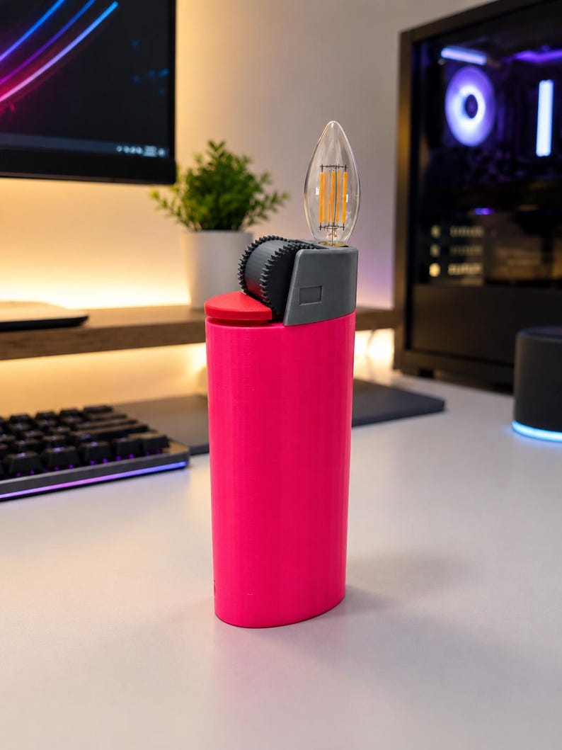 May include: A bright pink cylindrical lighter-shaped lamp featuring a grey top section with a textured ignition wheel and a clear, flame-shaped LED filament bulb extending from the top. The base is solid pink with a smooth, vertical finish.