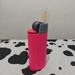 May include: A bright pink lighter-shaped object featuring a red top button, a dark gray textured ignition wheel, and a clear flame-shaped LED filament light bulb extending from the top where the flame would typically emerge.