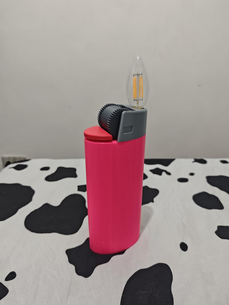 May include: A bright pink lighter-shaped object featuring a red top button, a dark gray textured ignition wheel, and a clear flame-shaped LED filament light bulb extending from the top where the flame would typically emerge.