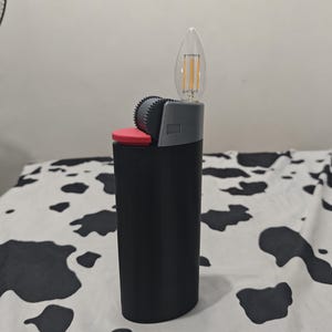 May include: A black lighter-shaped object featuring a red ignition button, a grey textured spark wheel, and a clear, flame-shaped LED filament light bulb extending from the top where the flame would typically emerge from a standard lighter.