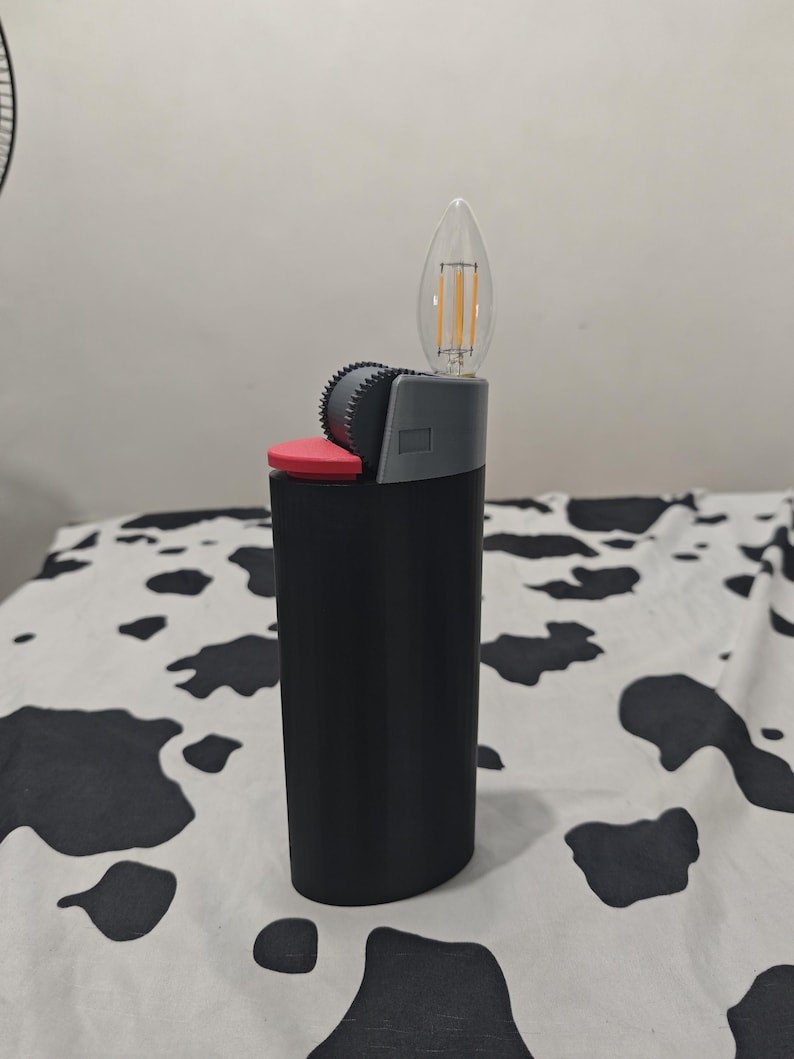 May include: A black lighter-shaped object featuring a red ignition button, a grey textured spark wheel, and a clear, flame-shaped LED filament light bulb extending from the top where the flame would typically emerge from a standard lighter.