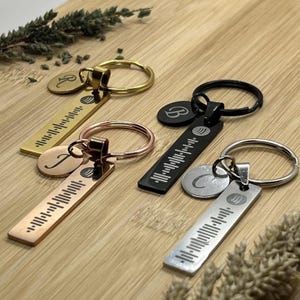 May include: Four keychains in gold, rose gold, black, and silver. Each keychain has a rectangular tag with a Spotify code and a circular tag with a letter. The keychains are on a wooden surface.