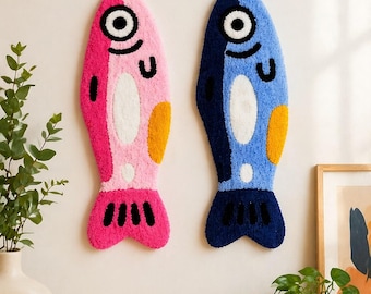 May include: Two tufted wall hangings shaped like fish. The left fish is pink and magenta with a white belly, yellow side patch, and black eye and fin details. The right fish is light and dark blue with a white belly, yellow side patch, and black eye and fin details.