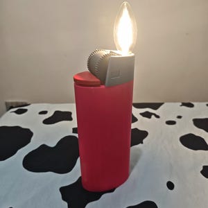 May include: A pink lighter-shaped lamp with a red top button and a grey ignition wheel. The top features a clear, flame-shaped light bulb that emits a warm glow, mimicking the appearance of a lit lighter in a vertical, cylindrical design.