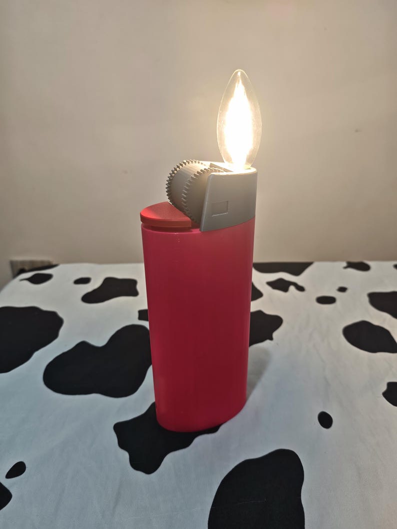 May include: A pink lighter-shaped lamp with a red top button and a grey ignition wheel. The top features a clear, flame-shaped light bulb that emits a warm glow, mimicking the appearance of a lit lighter in a vertical, cylindrical design.