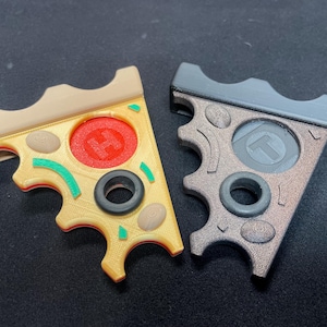 Puede incluir: Dos juguetes fidget con forma de porción de pizza. Uno es amarillo con detalles rojos, verdes y marrones, y el otro es gris y marrón. Ambos tienen un agujero circular en el centro y un diseño texturizado.