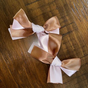 May include: Two hair bows crafted from smooth, shiny satin ribbon. Each bow features a layered design combining a metallic bronze-colored ribbon with a soft, pale pink ribbon. The ribbons are tied in a central knot, creating a classic bow shape.