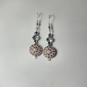 Op de afbeelding: Een paar zilveren oorhangers met witte en gouden knoopvormige bedels en heldere kristallen kralen.