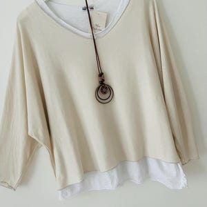 May include: A cream-coloured, layered top with a V-neck and a white undershirt. The top has a loose fit and long sleeves. A brown necklace with decorative rings and beads hangs in front of the shirt. The shirt is hanging on a white hanger.