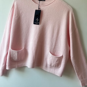 May include: A light pink long-sleeve jumper with a round neckline and two front patch pockets. The garment features a ribbed hem and cuffs, and includes a black brand tag hanging from the collar that displays the text AF ALPINO CASHMERE.