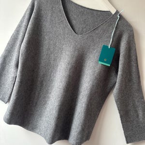 May include: A heather grey V-neck jumper with long sleeves, hanging on a white plastic hanger. A small, rectangular teal-coloured brand tag is attached to the neckline with a light blue string, featuring a circular logo at the bottom centre.