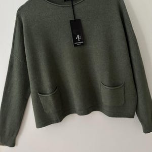 May include: An olive green sweater with a crew neck and two front pockets. The sweater has long sleeves and a ribbed hem and cuffs. A black tag with the brand logo hangs from the neckline. The sweater is hanging on a white hanger.