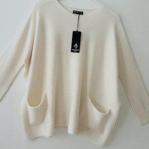 May include: An oversized cream-coloured sweater with long sleeves and two patch pockets. The sweater has a round neckline and is made of a soft, knitted fabric. The sweater has a tag that reads "4F ALENA FANTINI".
