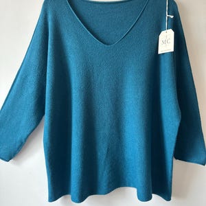 May include: A teal V-neck jumper with long sleeves and a relaxed fit. The garment features a textured knit fabric and includes a white rectangular paper tag attached to the neckline with a string, displaying the letters MC in a circular logo.