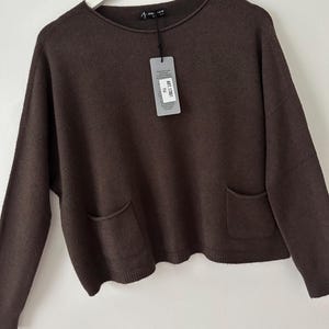 May include: A brown crew neck sweater with long sleeves and two front pockets. The garment has a tag attached to the neckline with the text "ART.17001 TU".