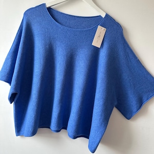 May include: A bright blue short-sleeved knit top with a wide, rounded neckline and a loose, boxy fit. The garment is displayed on a white hanger and features a small, rectangular beige brand tag attached to the neckline that reads New Notion.