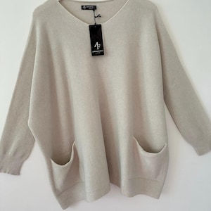 May include: A light beige knitted jumper with a V-neck and two patch pockets. The jumper has a ribbed texture and is made by the brand "4F Alpaca Fashion".