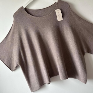 May include: A taupe-coloured knit jumper with a wide, loose fit, short sleeves, and a rounded neckline. The garment features a visible brand tag labelled "NEW COLLECTION" attached to the upper chest area, hanging from a white plastic hanger.