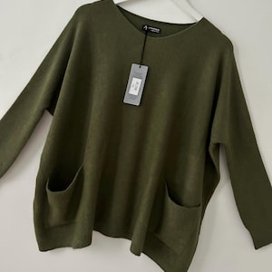 May include: An olive green sweater with a round neck and long sleeves. The sweater has two front pockets and a tag with the text "ART 2300". The garment is suspended from a white hanger.