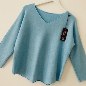 May include: A light blue long-sleeve jumper with a V-neckline and a relaxed fit. The garment is displayed on a white hanger and features a dark rectangular tag attached to the shoulder with the white text Y.Y. printed on the front surface.
