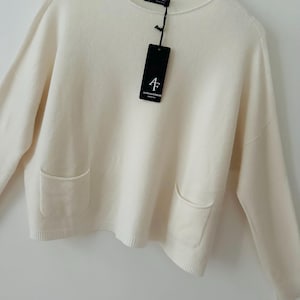 May include: Cream-coloured sweater with a crew neck and two front pockets. The long-sleeved knitwear has a relaxed fit and a tag with the brand name "ALPIN KNITWEAR". The sweater is hanging on a white hanger.